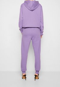 Fialová zkrácená mikina a odpovídající tepláky z měkké látky, s kapucí, žebrovanými manžetami a tapered nohavicemi. Nošeno s černými sandály na podpatku.