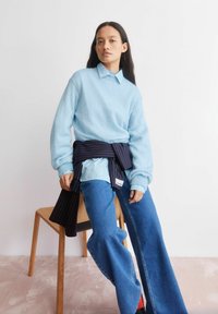 Hellblauer Strickpullover mit einem Hemd mit Kragen darunter, kombiniert mit hoch taillierten blauen Jeans und einem marineblauen, geringelten Wickeltuch, das um die Taille gebunden ist.