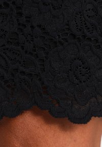 Svart blommönstrad spetsduk med intrikata mönster över brun hud, vilket visar detaljerad textur och scallopkant.