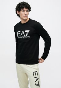 Férfi fekete hosszú ujjú felsőben, rajta fehér "EA7 Emporio Armani" logóval, és krémszínű nadrágban, melynek combján fekete "EA7" logó látható, fehér háttér előtt áll.