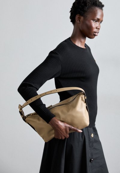 Sac à épaule en cuir beige avec une texture lisse et une bandoulière ajustable. Le mannequin porte un pull noir à côtes et une jupe noire.