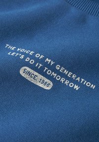 Tissu bleu avec texte blanc disant « La voix de ma génération, faisons-le demain » et « Depuis 1986 » dans un rectangle arrondi.