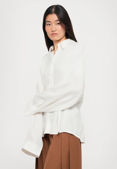 Young woman with straight dark hair wearing an oversized white button-up shirt and brown pleated trousers facing front against white background.