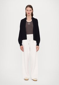 Svart cardigan med droppede skuldre over en brun tanktop med "MM." Hvite vidbenede bukser og beige slip-on sko fullfører antrekket.