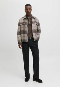 Muž v károvaném béžovo-hnědém saku, hnědém svetr s zipem, černých kalhotách s opaskem a černých botách, stojící s jednou rukou v kapse.