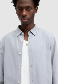 Lichtgrijze button-up shirt in textuurstof, met een puntkraag en lichte knopen, gelaagd over een witte geribbelde tanktop.