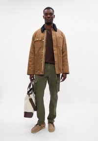 Brun bomuldsjakke med sort krave, brun t-shirt, grønne cargobukser med lommer, beige sko og en beige taske med brune detaljer.