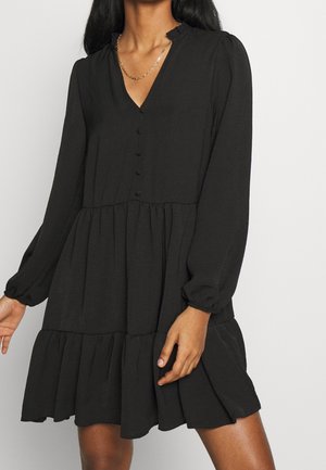 Žena v dlouhých černých šatech na knoflíky s vrstvenou sukní a zlatým řetízkovým náhrdelníkem stojí před jednobarevným pozadím.