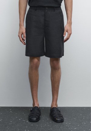 Pantaloni shorts neri in lino con vestibilità comoda, caratterizzati da un fronte piatto e tasche laterali, abbinati a sandali neri con un design alla schiava.