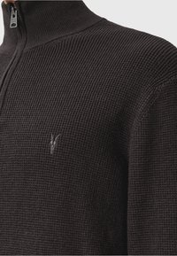 Close-up van een donker gebreide trui met een kwart-rits kraag en een klein geborduurd dierenlogo op de borst.