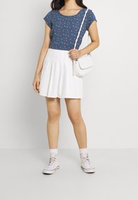 Tryckt blå topp med kort ärm och små blommönster, tillsammans med en vit plisserad kjol och vit handväska. Avslutats med vita sneakers.