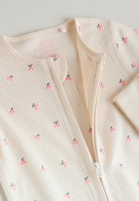 Beige ribbattua vauvan body, jossa on edessä vetoketju ja pieniä vaaleanpunaisia kirsikkakuvioita kankaalla.