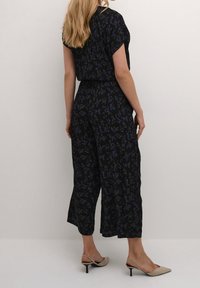 Zwarte jumpsuit met bloemenpatroon in blauw en groen, korte mouwen, losse pasvorm, cropped broekspijpen en elastische tailleband. Gestyled met beige hakken.