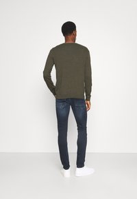Maglietta verde a maniche lunghe con scollo rotondo, abbinata a jeans slim-fit blu scuro e sneakers bianche. Il modello è di spalle rispetto alla fotocamera.