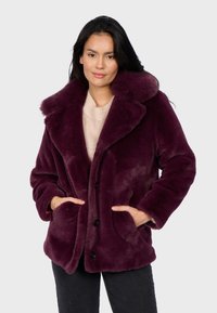 Mujer con cabello largo y oscuro que lleva un abrigo de piel sintética morado profundo sobre un suéter beige, con las manos en los bolsillos del abrigo, de pie contra un fondo claro.