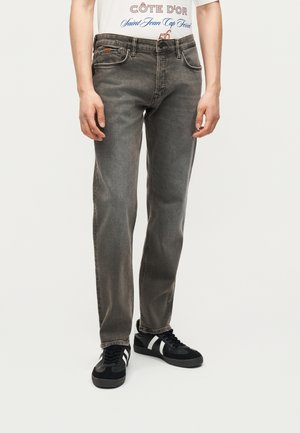 Férfi szürke egyenes szárú farmerben, fekete, fehér csíkos tornacipőben és fehér grafikás pólóban, egyszínű háttér előtt áll.