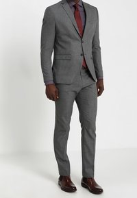 Grau maßgeschneiderter Anzug mit dezentem Muster, kombiniert mit einer bordeauxroten Krawatte. Dazu ein kariertes Hemd und braune Leder-Herrenschuhe.