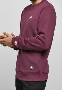 Hombre con un jersey de cuello redondo color burdeos con los logos de Starter Black Label en el pecho, el puño de la manga y el dobladillo, de pie con las manos entrelazadas.