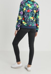 Mujer vestida con jeans ajustados oscuros, zapatillas blancas y una chaqueta azul marino con estampado floral colorido, de pie con una pierna doblada.