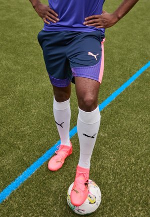Personne debout sur du gazon artificiel, un pied sur un ballon de football, portant un short violet, des chaussettes blanches et des chaussures de football rose vif.