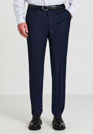 Homme portant un pantalon habillé à carreaux bleu marine avec une ceinture noire, des chaussures en cuir noires, une chemise blanche, une main dans la poche, se tenant sur un fond blanc.