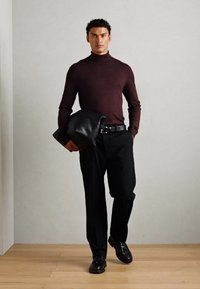 Bordeaux turtleneck trui, zwarte broek, zwarte riem en schoenen. Houdt een zwarte leren clutch vast. Effen achtergrond met houten vloer.