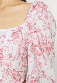 Blouse blanche avec un motif floral rouge, dotée d'un col carré et de manches bouffantes. Tissu léger et texturé avec des fronces aux coutures des épaules.