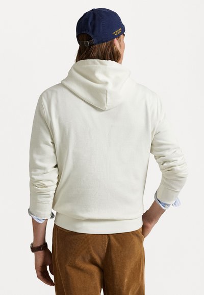 Hvit hettegenser med en noe oversized passform, med en stor hette og ribbestrikkede mansjetter. Bres over brune kordfløyelsbukser, som viser en lagdelt skjorte under.