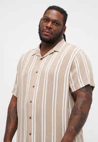 Camisa de manga corta a rayas en beige y blanco, con cuello, cierre de botones en la parte delantera y un ajuste relajado. Tejido texturizado con costuras visibles.
