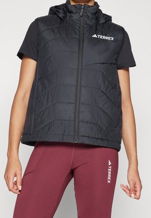 Osoba nosząca czarną bezrękawnikową pikowaną kamizelkę z kapturem i zamkiem błyskawicznym, czarną koszulkę z krótkim rękawem oraz bordowe getry z logo Terrex.