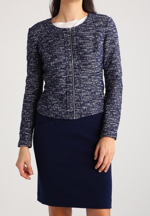 Femme portant une veste zippée texturée bleu marine sur un haut blanc, associée à une jupe bleu marine longueur genou sur un fond uni.