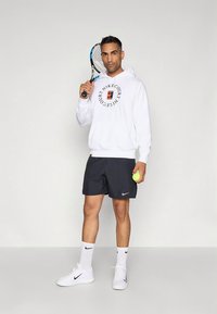 Sweatshirt branco com logótipo circular 'Nike Court', calções pretos, sapatos brancos, segurando uma raquete de ténis e uma bola de ténis verde, fundo neutro.