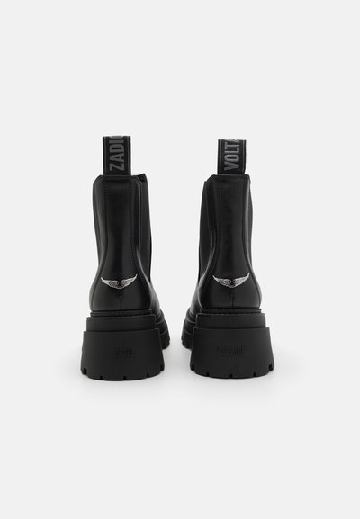 Botas de cuero negras hasta el tobillo con paneles laterales elásticos, suelas de goma gruesas, acentos metálicos en las alas y tiradores etiquetados como "ZADIG" y "VOLTAIRE."