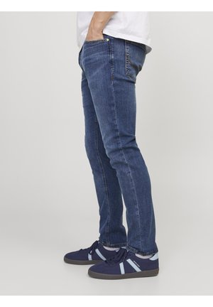 Personne portant un jean bleu slim et des baskets bleu marine avec des bandes bleu clair, debout, une main dans la poche, sur un fond uni.