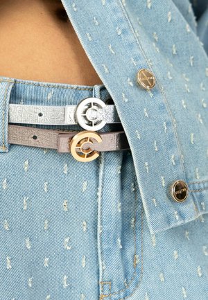 Dva remena u srebrnoj i sivoj boji s okruglim metalnim kopčama, nošeni preko traper košulje svijetloplave tkanine s malim bijelim uzorcima teksture.