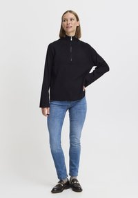 Maglietta nera a maniche lunghe con colletto alto e corta zip, abbinata a jeans slim blu chiaro e mocassini neri, in piedi davanti a uno sfondo neutro.