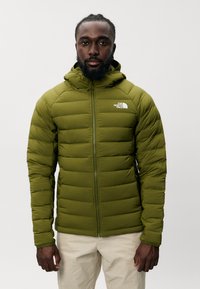 Giacca puffer verde oliva con chiusura a zip e cappuccio, caratterizzata da sezioni trapuntate orizzontali e un piccolo logo sul lato sinistro del petto.