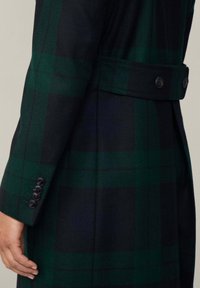 Tmavozelený a námornícky károvaný plášť s rukávmi a zadnou časťou, štyri čierne gombíky na manžete a dva gombíky na opasku na chrbte, ruka uvoľnená pri tele.