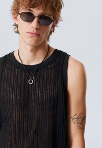 Haut sans manches noir avec des rayures transparentes verticales, porté avec des lunettes de soleil ovales et un collier noir avec un pendentif circulaire. Un tatouage sur le bras est visible.
