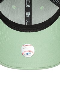 Boné de tecido verde com ponta preta, apresentando um autocolante com o logótipo de uma equipa de beisebol na parte inferior. Tecido texturizado com detalhes de costura visíveis.