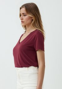T-shirt bordeaux a maniche corte con scollatura a V, realizzato in tessuto morbido, indossato con jeans bianchi a vita alta, evidenziando uno stile casual.