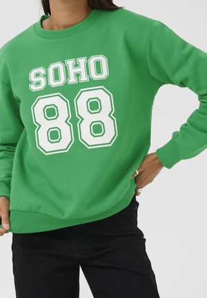 Osoba w zielonej bluzie z białym napisem "SOHO 88", w połączeniu z czarnymi spodniami, pozująca z jedną ręką na biodrze.