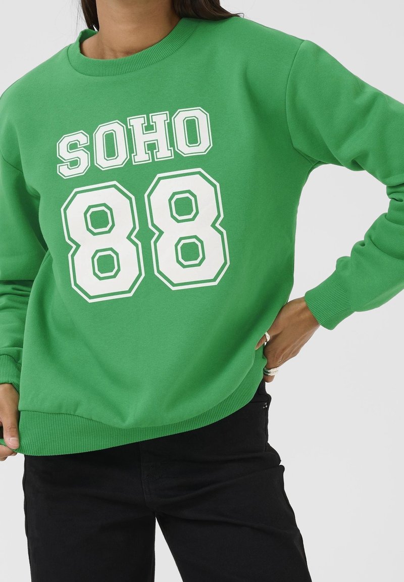 Personne portant un sweat-shirt vert avec l'inscription blanche "SOHO 88", associé à un pantalon noir, posant avec une main sur la hanche.