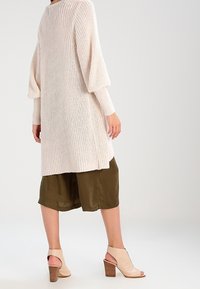 Pitkä, vaalea beige neulecardigan, jossa on ribbireunat, yhdistettyinä oliivinvihreisiin shortseihin. Korostettu avonaisilla beigeillä koroilla.