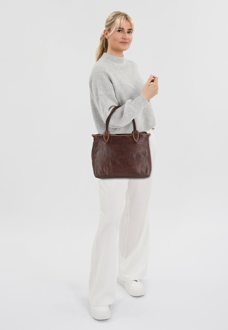 Brown leren totebag met twee handvatten, met een textuurafwerking en decoratieve accenten op de bovenrand. Model draagt een grijze trui en witte broek.