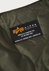 Étiquette en tissu noir sur matériau matelassé vert olive avec le logo "Alpha Industries" et le texte : Knoxville, Tennessee, États-Unis ; établi en octobre 1959 ; entrepreneur du Département de la Défense des États-Unis.