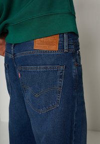Mörkblå denimjeans med rak skärning, som har en brun läderpatch på baksidan, gula sömmar och en klassisk femficksdesign.