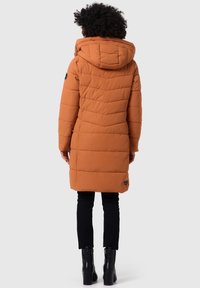Lange oranje pufferjas met capuchon, voorzien van een gewatteerd ontwerp en een logo-opdruk op de mouw. Draagt over zwarte broeken en laarzen.