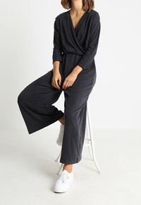 Jumpsuit gris foncé avec un design à devant drapé, manches longues, jambes larges et une coupe confortable ; associé à des baskets blanches.