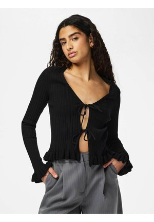 Vrouw met krullend haar, gekleed in een zwart geribd vestje geknoopt aan de voorkant en grijze broek met krijtstrepen, handen in de zakken, neutrale uitdrukking.